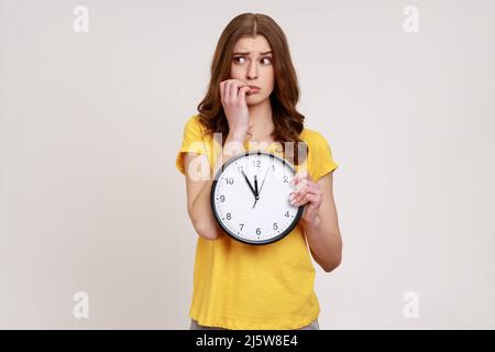 Ungeduldig nervös Teenager-Mädchen mit welligen Haaren in gelben T-Shirt sitzen beißen ihre Nägel mit großen Wanduhr, Frist, afraids zu spät zu sein. Innenaufnahme des Studios isoliert auf grauem Hintergrund. Stockfoto
