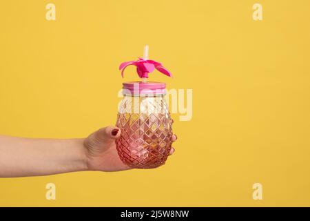 Nahaufnahme der Hand einer Frau, die einen Becher aus rosafarbenem Glas mit Strohhalm für einen Cocktail oder Smoothie hält. Innenaufnahme des Studios isoliert auf gelbem Hintergrund. Stockfoto
