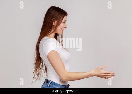 Lassen Sie mich mich mich vorstellen. Seitenansicht Porträt einer freundlichen Frau, die Hand zum Händeschütteln gibt, die Gäste mit einem dutzenden Lächeln begrüßt und ein weißes T-Shirt trägt. Innenaufnahme des Studios isoliert auf grauem Hintergrund. Stockfoto