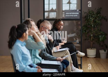 Afrikanische junge Frau liest einen Vertrag, während sie mit anderen Menschen im Flur im Büro sitzt und auf ihr Vorstellungsgespräch wartet Stockfoto