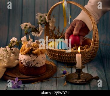 Osterkuchen, ein Korb mit farbigen Eiern, ein bronzener Kerzenständer mit brennender Kerze, eine Hand mit einem Ei und Frühlingsblumen mit einem verschwommenen Backgroun Stockfoto