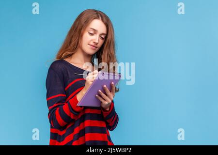 Positive Frau trägt gestreiften lässigen Stil Pullover, schriftlich in Veranstalter, machen zu tun Liste, Designer Skizze zeichnen oder Notizen. Innenaufnahme des Studios isoliert auf blauem Hintergrund. Stockfoto