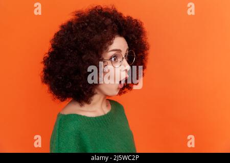 Profilportrait einer schockierten Frau mit Afro-Frisur in grün lässiger Pullover mit offenem Mund stehend, wegblickend, sieht etwas Überraschendes. Innenaufnahme des Studios isoliert auf orangefarbenem Hintergrund. Stockfoto