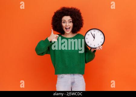 Porträt einer glücklichen, aufgeregten Frau mit Afro-Frisur, die einen grünen Pullover im lässigen Stil trägt, der mit einer Wanduhr in den Händen steht und den Daumen nach oben zeigt. Innenaufnahme des Studios isoliert auf orangefarbenem Hintergrund. Stockfoto