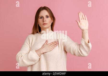 Portrait einer ehrlichen blonden Frau, die mit einem Promise-Handzeichen steht, Handfläche nach oben gestikuliert, Versprechen gibt, Treue verspricht, weißen Pullover trägt. Innenaufnahme des Studios isoliert auf rosa Hintergrund. Stockfoto