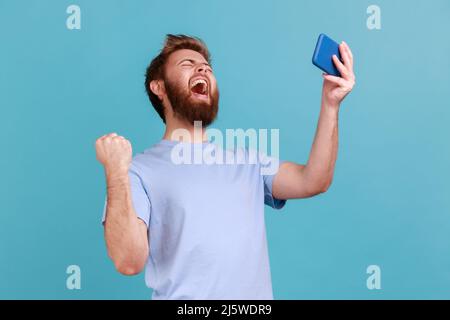 Porträt von extrem glücklich gut aussehend bärtigen Mann Gamer spielen Videospiel auf dem Handy, feiert den Sieg, komplette Ebene. Innenaufnahme des Studios isoliert auf blauem Hintergrund. Stockfoto