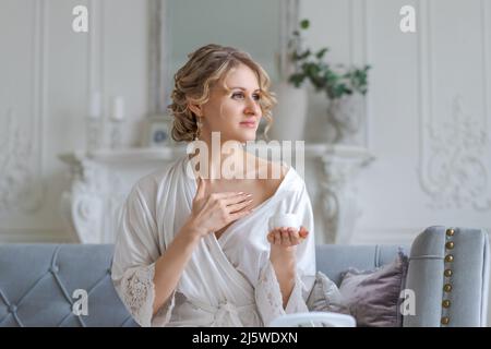 Hübsche europäische Frau trägt Gesichtscreme auf ihr Gesicht hält Glas Kosmetik-Produkt kümmert sich um ihre Haut und Blume, Posen auf bequemen Sofa in Seidengewand, während im Wohnzimmer zu Hause am Morgen sitzen Stockfoto