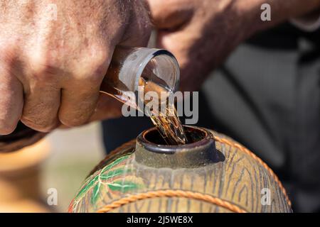 Mann gießt Wein aus der Glasflasche in ein Keramikfass. Hand und Wein, Nahaufnahme Stockfoto