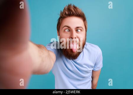 Porträt eines hübschen, kindisch bärtigen Bloggers, der ein Sichttelefon macht, die Zunge zeigt, kindhaftes Verhalten demonstriert, POV. Innenaufnahme des Studios isoliert auf blauem Hintergrund. Stockfoto