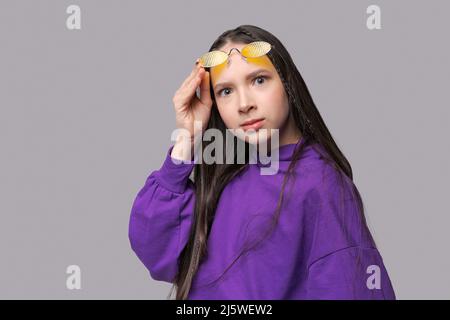 Portrait niedlichen Teenager-Mädchen in lila Sweatshirt und gelbe Brille posiert im Studio auf einem grauen Hintergrund. Kaukasische Mädchen raotae vor der Kamera Stockfoto