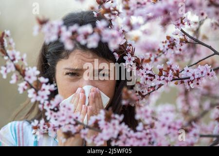 Ängstlich junge Frau, die vor blühendem Baum mit Serviette die Nase wischend. Frühjahrsallergie-Angriffskonzept Stockfoto