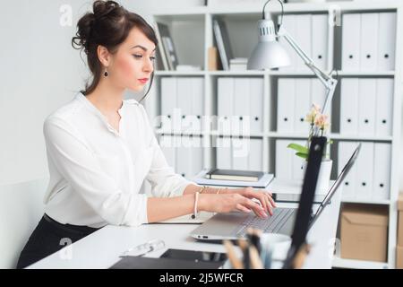 Porträt einer jungen Büroangestellerin, die im Büro an ihrem Arbeitsplatz sitzt, tippt, aufmerksam auf den Laptop-Bildschirm schaut Stockfoto