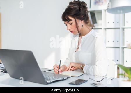 Junge weibliche Angestellte, die im modernen Büro sitzt und Notizen in ihrem Notizblock macht. Mitarbeiter, der mit verschiedenen digitalen Geräten am Arbeitstisch arbeitet Stockfoto