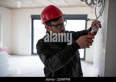 Seriöse junge männliche Elektriker mit Helm hält schneiden elektrische Drähte mit einer Zange. Stockfoto