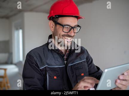Junger lächelnder männlicher Ingenieur in Schutzuniform mit Helm, der in der Fabrik Tablet verwendet. Stockfoto