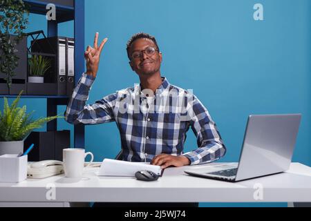 Fröhliche, fröhliche afroamerikanische Büroangestellte, die mit den Fingern eine Siegesfriedensgeste macht, während sie die Kamera anschaut. Lächelnder, fröhlicher Mann, der im Büro mit der Hand das Siegersymbol macht. Stockfoto