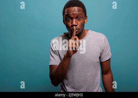 Junger Mann, der eine Geste mit dem Finger über den Lippen tut, um über vertraulichen Tratsch geheim zu halten. Geheimnisvolle Person, die ein Stummschaltungssymbol zeigt, um bei privaten Gesprächen still und leise zu sein. Stockfoto