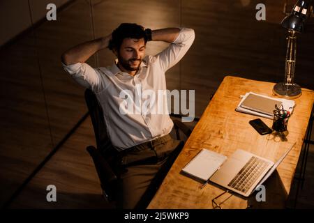 Weißer bärtiger Mann, der lächelte, als er abends im Büro mit einem Laptop arbeitete Stockfoto