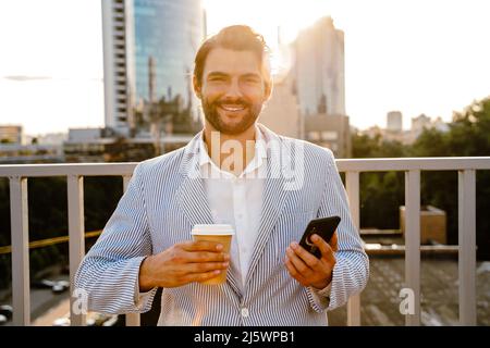 Junger Mann lächelt und benutzt Handy, während er Kaffee im Freien trinkt Stockfoto