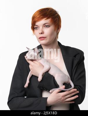 Junge Frau mit kurzen Haaren hält in den Händen Sleeping Sphynx Hairless Cat blauen Nerz und weiße Farbe. Schöne rothaarige Frau in schwarzer Jacke gekleidet Stockfoto