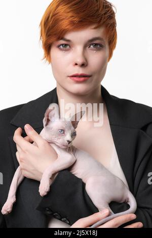Serious junge Frau in schwarzer Jacke hält in den Händen Canadian Sphynx Cat. Hübsche Rothaarige Frau, 25 Jahre alt, mit kurzen Haaren und Blick auf die Kamera Stockfoto