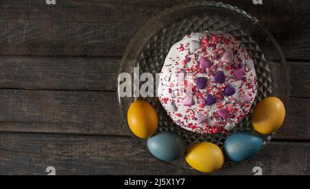 Osterkuchen in einer transparenten Schale, um bemalte Eier in den Farben der Flagge der Ukraine. Gelb und blau (Cyan). Auf Holzstruktur. Konzept: Essen, h Stockfoto