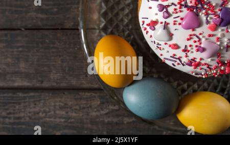 Osterkuchen in einer transparenten Schale, um bemalte Eier in den Farben der Flagge der Ukraine. Gelb und blau (Cyan). Auf Holzstruktur. Konzept: Essen, h Stockfoto