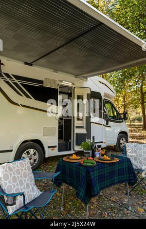 Wohnmobil auf dem Campingplatz mit Tisch und Stühlen, die außerhalb des Fahrzeugs aufgestellt und zum Essen und Trinken bereit sind. Stockfoto