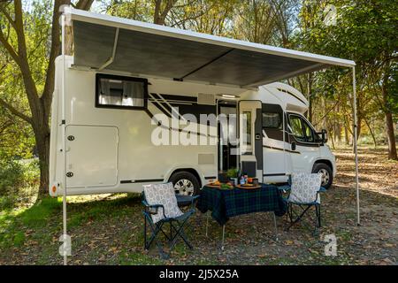 Wohnmobil auf dem Campingplatz mit Tisch und Stühlen, die außerhalb des Fahrzeugs aufgestellt und zum Essen und Trinken bereit sind. Stockfoto