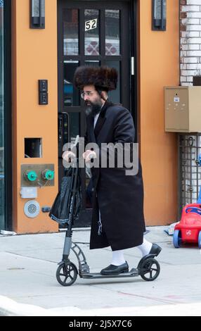 Auf Passah und in langen weißen Strümpfen und einem Shtreimel Pelzhut, kauft und reitet ein orthodoxer jüdischer Mann seinen Roller. An der Lee Ave in Williamsburg NYC. Stockfoto