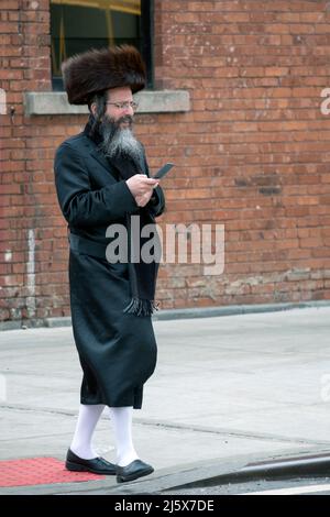 Auf Passah und in langen weißen Strümpfen und einem Shtreimel-Pelzhut überprüft ein orthodoxer jüdischer Mann sein Handy. An der Bedford Ave in Wiliamburg, NYC. Stockfoto