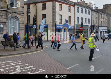 Osterprozession mit einem großen Kreuz und einem Banner, das die Straße an der Ampel in Kendal, Cumbria, Großbritannien, überquert, wobei ein Mann in einer Hi-viz-Jacke anschaut Stockfoto