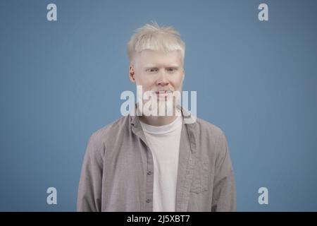 Porträt eines jungen Albino-Mannes mit weißem Haar, blasser Haut und blauen Augen, der vor blauem Hintergrund auf die Kamera blickt Stockfoto