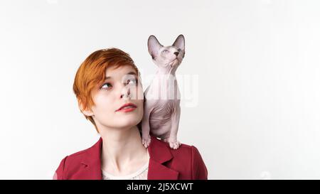 Schöne Hipster Rothaarige junge Frau mit kurzen Haaren aufschauend, gekleidet in rote Jacke und verspielte Sphynx Kätzchen sitzt auf ihrer Schulter. Studioaufnahme Stockfoto