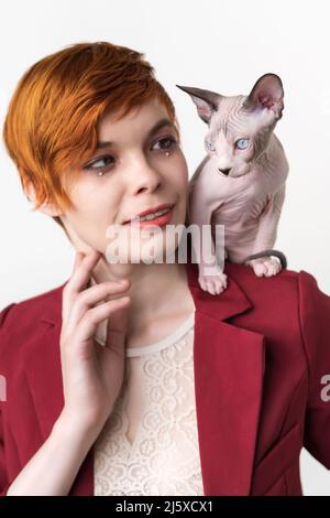 Porträt einer jungen Hipster-Rotschopf-Frau mit kurzen Haaren, in roter Jacke und verspielter Sphynx Cat auf ihrer Schulter. Studio auf Weiß aufgenommen Stockfoto