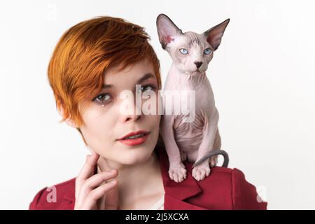 Porträt einer schönen rothaarigen jungen Frau mit kurzen Haaren, in roter Jacke gekleidet und verspielte Katze, die auf ihrer Schulter sitzt. Hipster Frau suchen Stockfoto