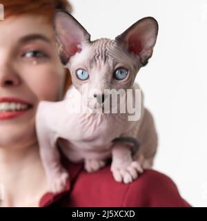 Sphynx Hairless Cat blickt auf die Kamera, die auf der Schulter einer lächelnden, rothaarigen jungen Frau in roter Jacke sitzt. Vorderansicht, Studioaufnahme auf Weiß Stockfoto