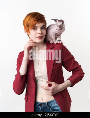 Porträt einer jungen Hipster-Rotschopf-Frau mit kurzen Haaren, bekleidet mit roter Jacke, blauer Jeans und verspielter Sphynx Cat, die auf ihrer Schulter sitzt. Vorderansicht Stockfoto