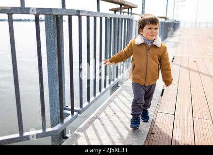 Ein kleiner fröhlicher cooler Junge in stylischer, modischer, warmer Kleidung geht entlang eines weiten, leeren Piers mit einem alten rostigen grauen Zaun in der Nähe des ruhigen, ruhigen, großen Black S Stockfoto