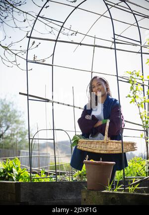 Portrait lächelnde junge Frau, die unter einem Gitter im sonnigen Garten gärtelt Stockfoto