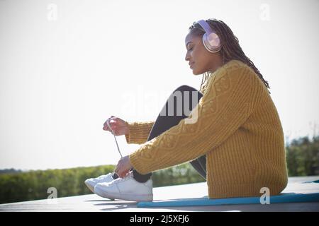 Junge Frau mit Kopfhörern, die im Freien Schuhe binden Stockfoto