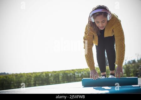 Junge Frau mit Kopfhörern, die auf der sonnigen Terrasse eine Yogamatte hochrollt Stockfoto