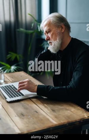 Die vertikale Aufnahme eines müden grauhaarigen, reifen Geschäftsmanns, der am Arbeitsplatz sitzt, hat starke Nackenschmerzen und reibt ihn, um Muskelverspannungen zu lindern. Stockfoto