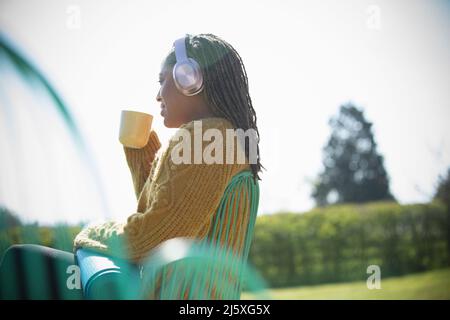 Junge Frau mit Kopfhörern und Yogamatte, die draußen Kaffee trinkt Stockfoto