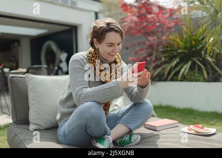 Lächelnde junge Frau, die Smartphone auf dem Loungesessel auf der Terrasse benutzt Stockfoto