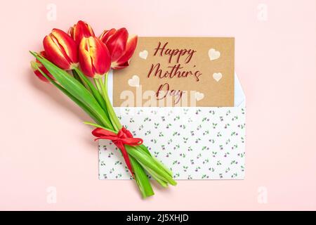 Blumenstrauß aus roten Tulpen, Text Happy Mother's Day auf Karte im Umschlag auf rosa Hintergrund Draufsicht Flat Lay Holiday Grußkarte. Stockfoto