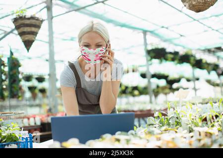 Weibliche Gartengeschäft Inhaberin in Gesichtsmaske Gespräch am Telefon am Laptop Stockfoto