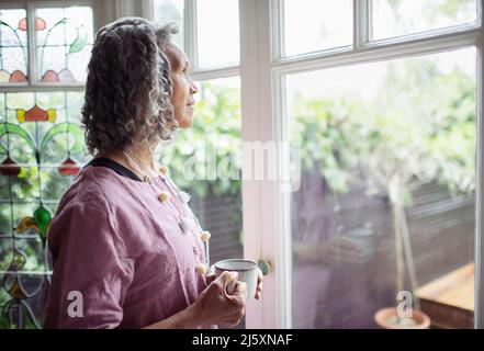 Nachdenkliche ältere Frau, die am Fenster Tee trinkt Stockfoto