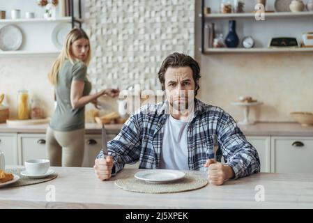 Wütend hungrig unzufrieden tausendjährigen kaukasischen Mann mit Stoppeln ignorieren Frau und warten auf Essen in der Küche Stockfoto