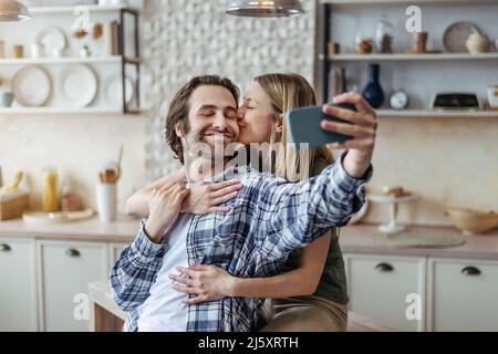 Lächelnde junge europäische Frau umarmt und küsst ihren Mann mit Stoppeln, Kerl macht Selfie auf dem Smartphone Stockfoto